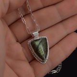 green arrow shaped pendant featuring a green labradorite stone, hanging on a paperclip style chain shown in hand for size