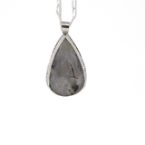 Teardrop shaped silver labradorite pendant with flashes of rainbow colors set in sterling silver, with a hand textured decorative border surrounding the stone and hanging from a paperclip style chain